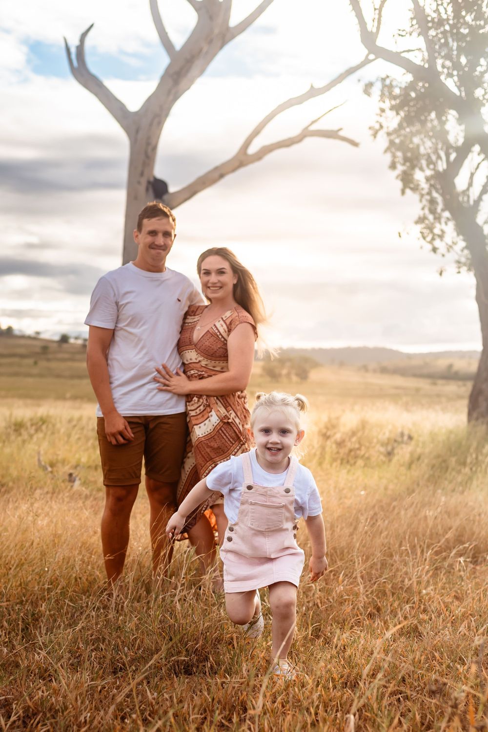 A happy family poses in a sunlit field with their young child running in the foreground.