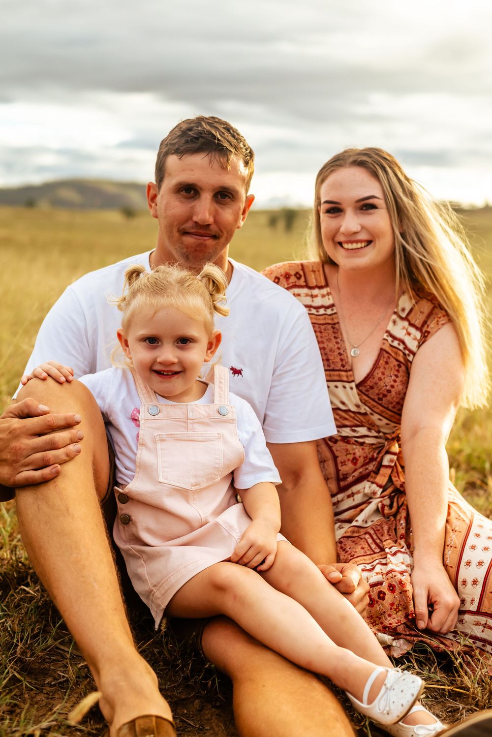 Family sitting together in a grassy field during golden hour with child in pink overalls.