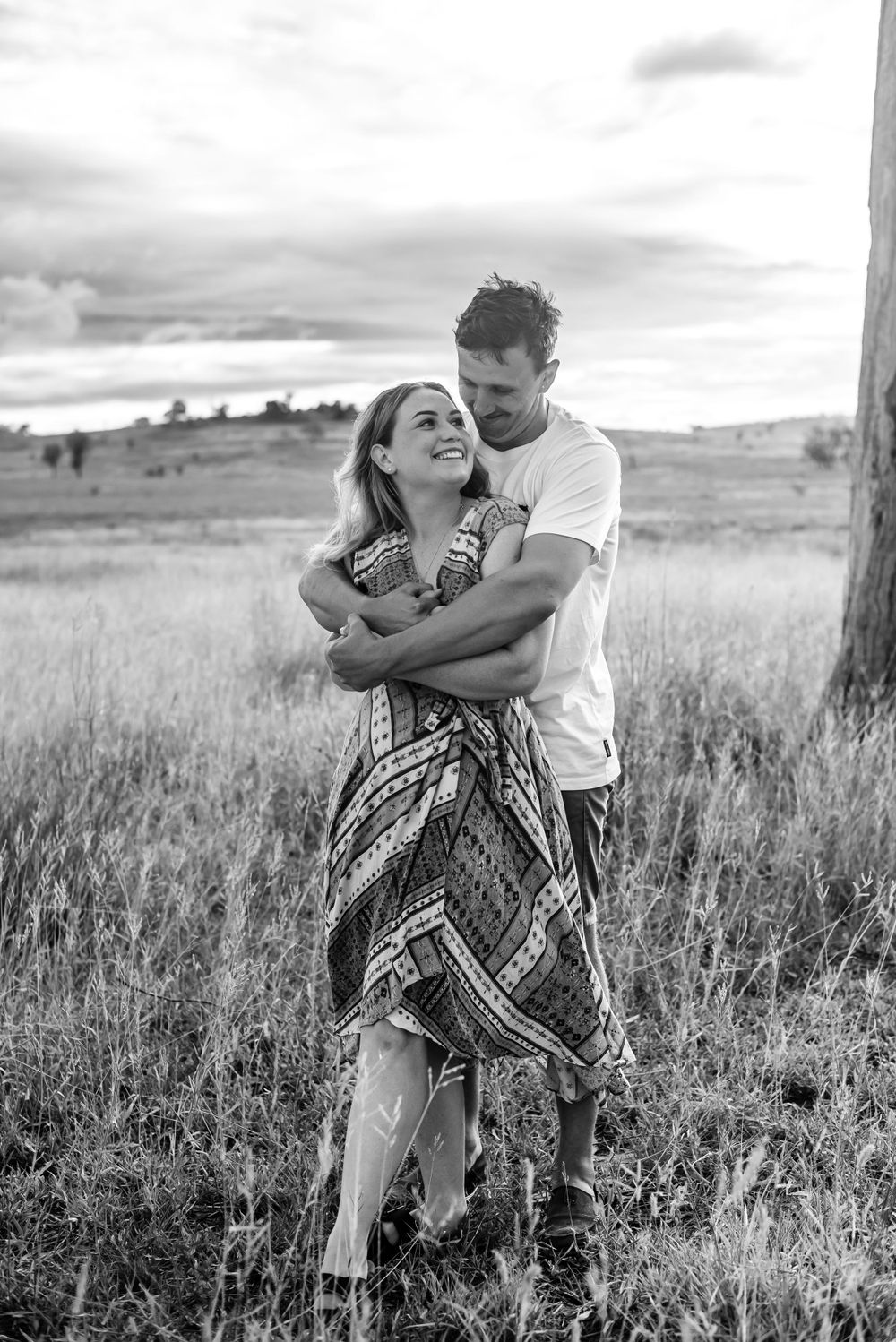 A romantic black and white photo of a couple embracing in a grassy field with cloudy skies in the background.