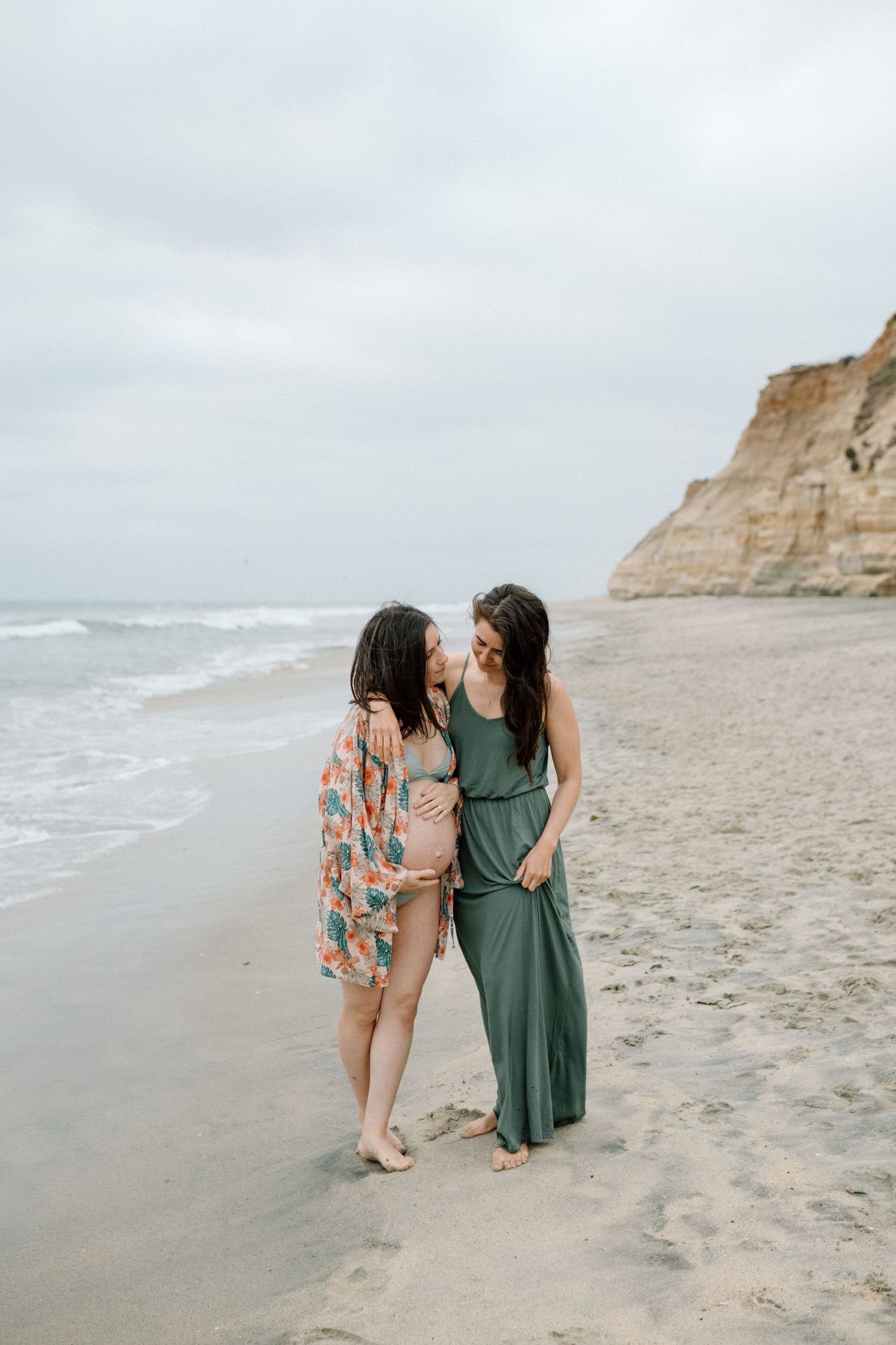 Two women in flowing dresses sharing a moment on a beach near dramatic coastal cliffs during maternity photoshoot in San Diego.