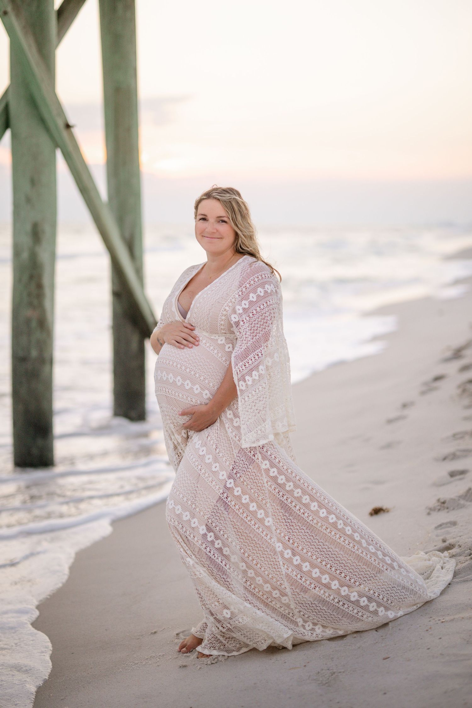Beachside maternity portrait in a lace gown next to wooden pier pillars.