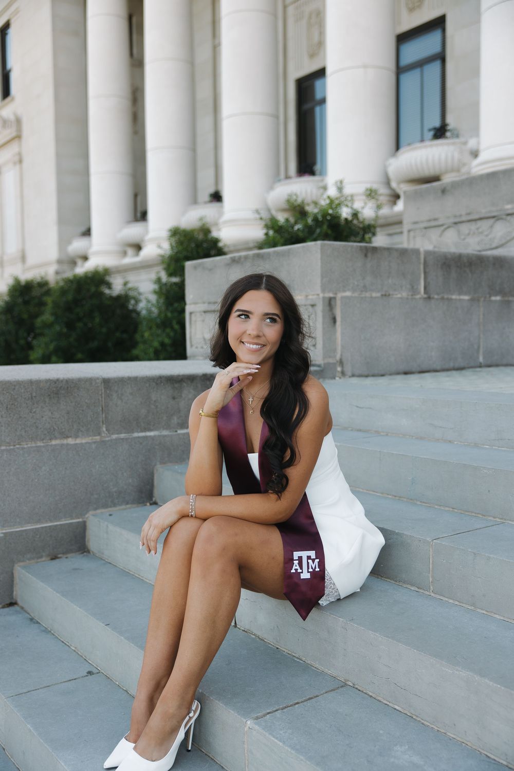 Texas A&M graduate smiling on stone steps wearing white dress and maroon Aggie sash.