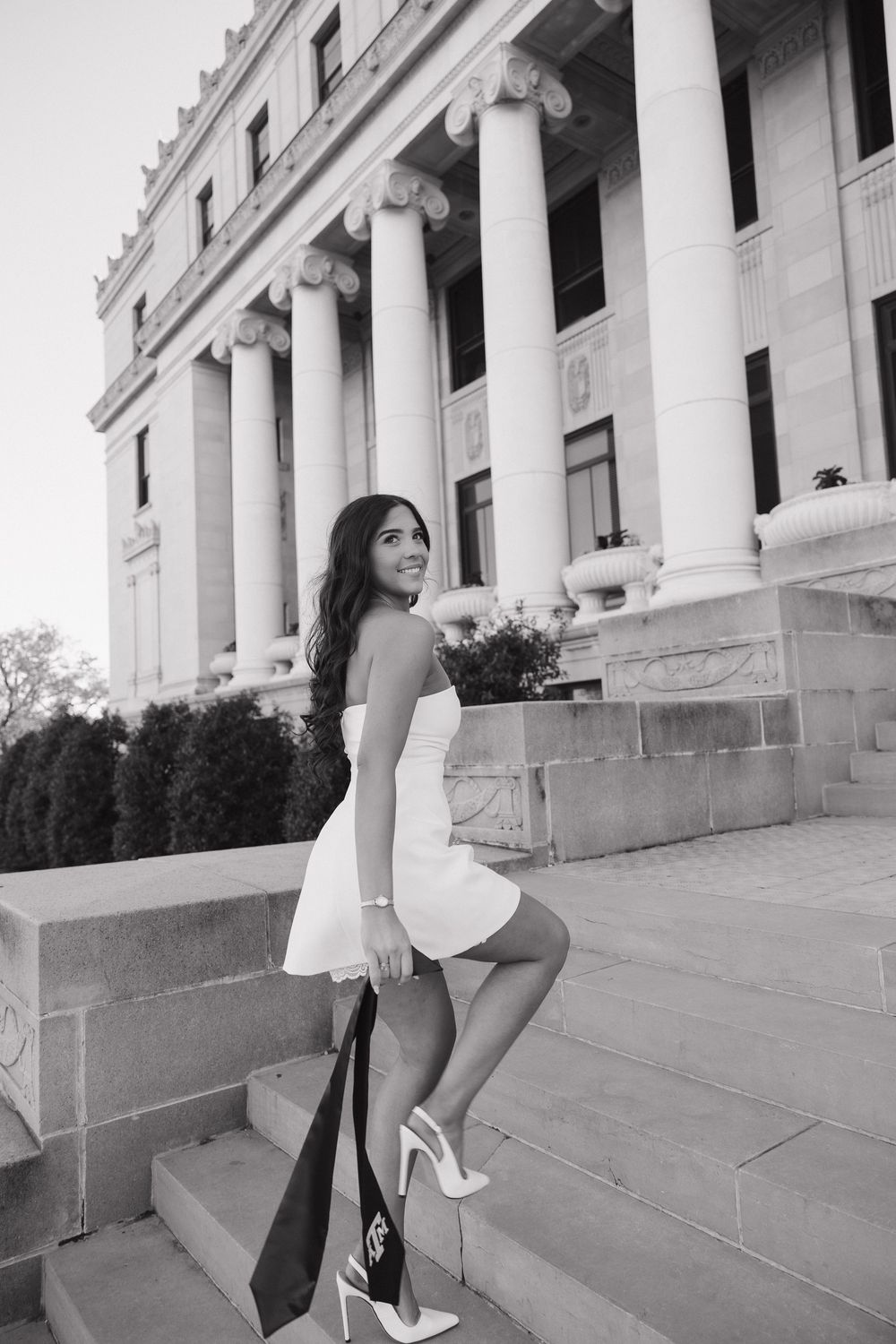 Black and white photo of elegant woman in white dress and heels on steps of neoclassical building.