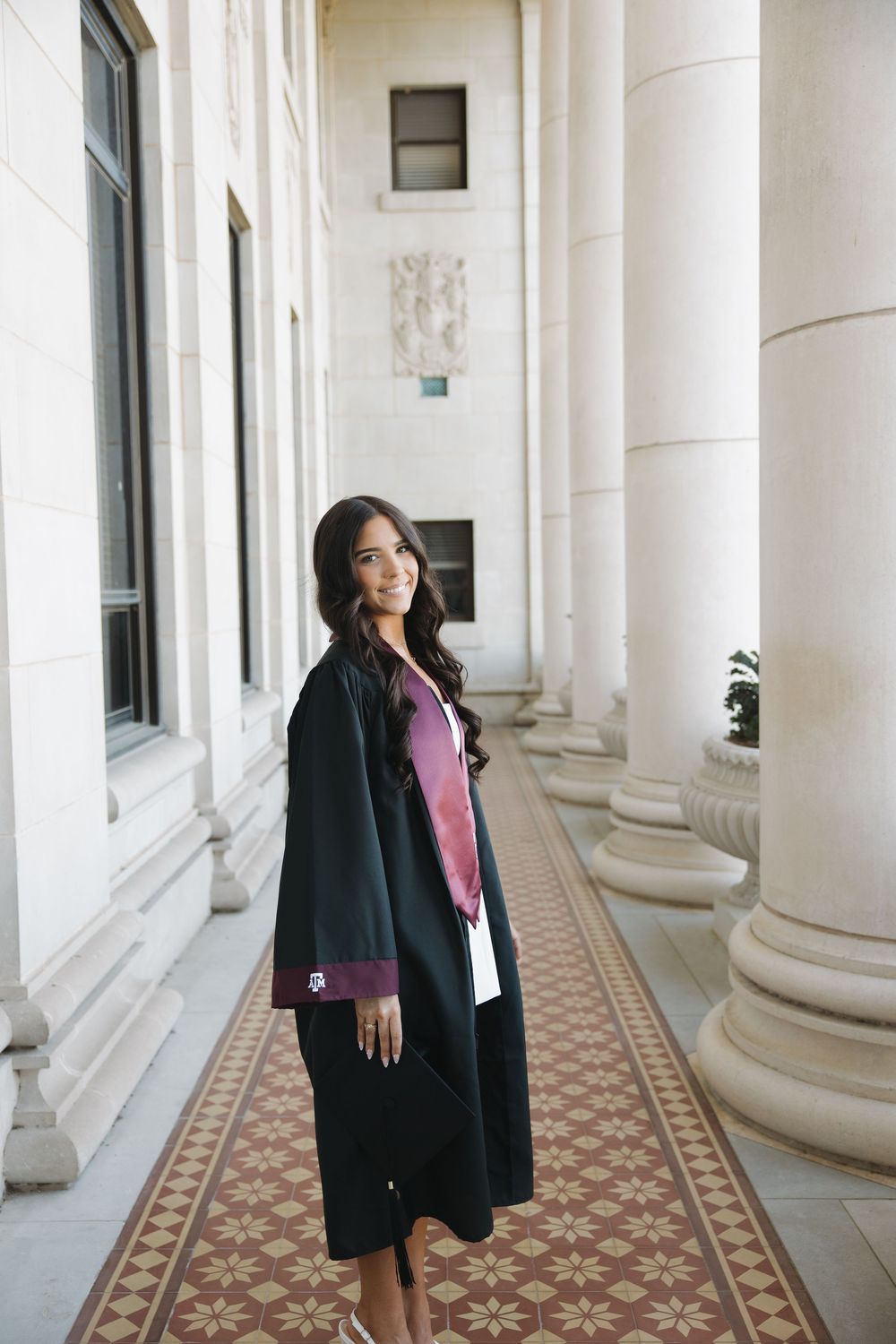 Young woman in black graduation gown with maroon trim posing in elegant columned hallway.