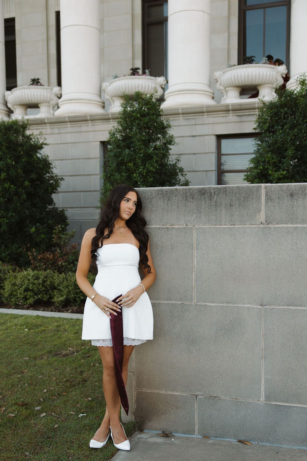 Young woman in white strapless dress and heels posing against stone wall outside elegant building with columns.