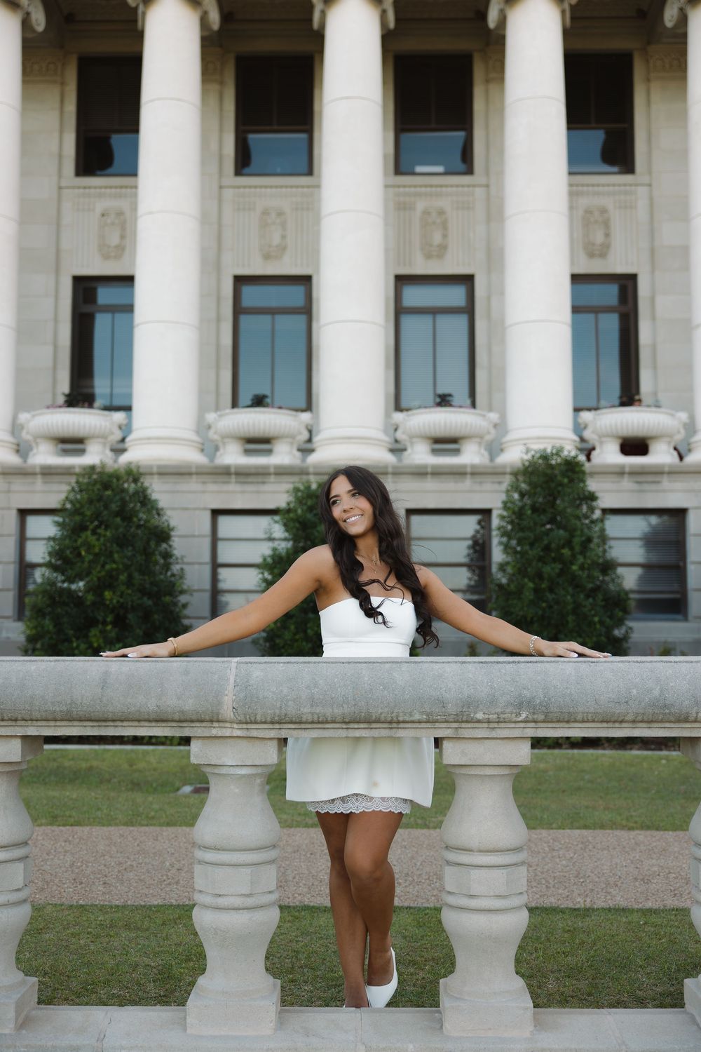 Young woman in white dress smiling, leaning on stone balustrade before a neoclassical building with tall columns.