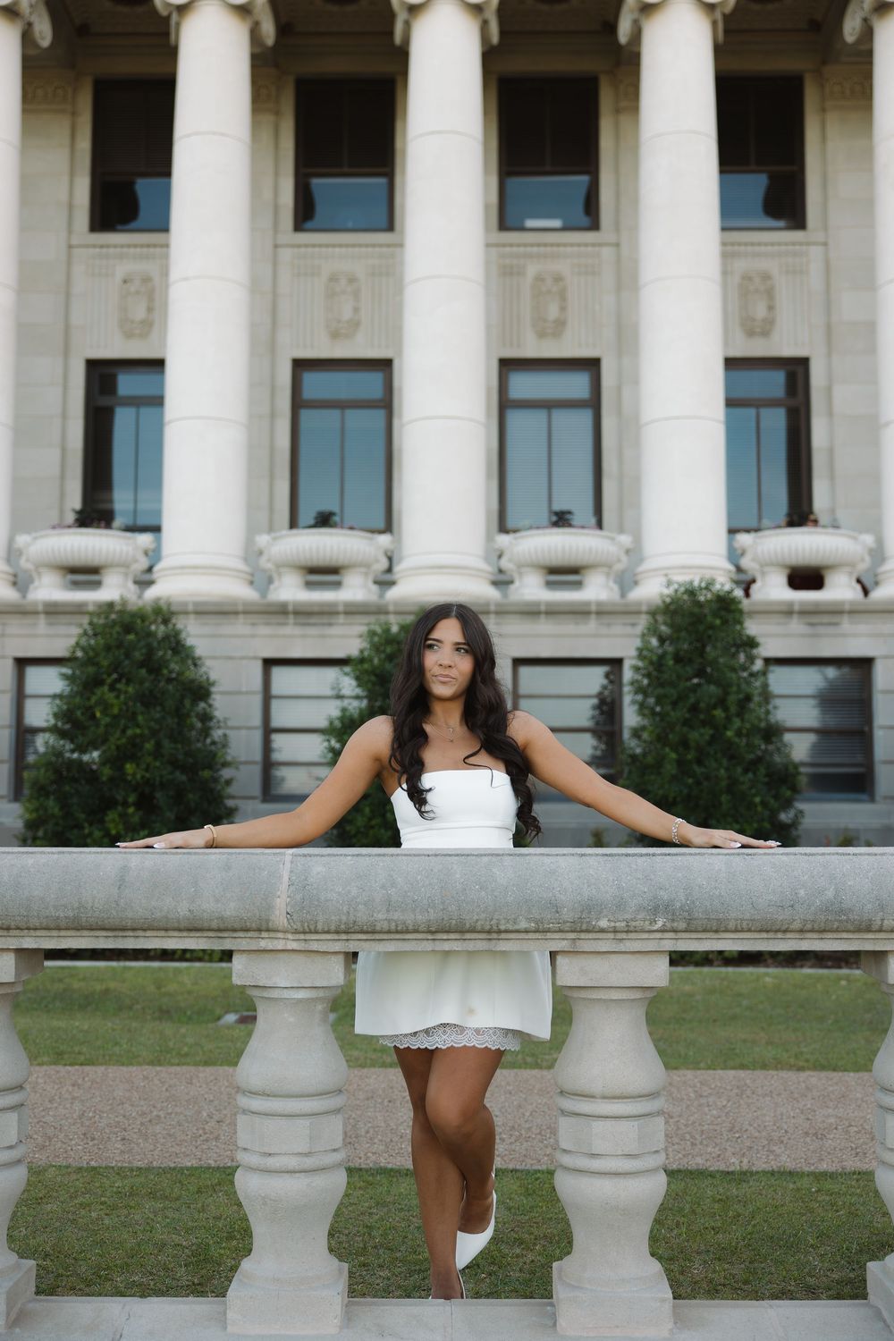Elegant woman in white mini dress posing confidently at stone railing outside a grand neoclassical building.