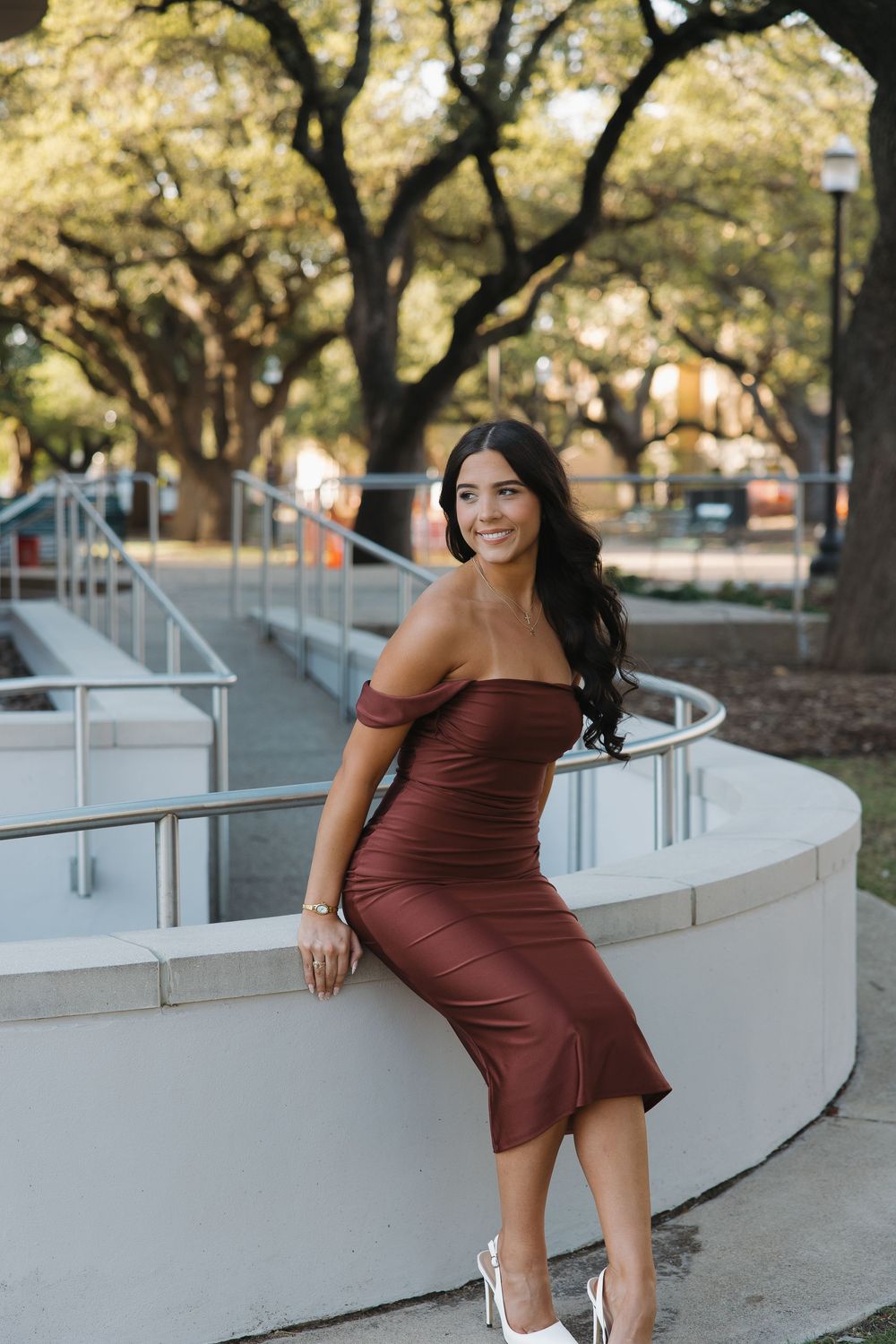 Young woman in brown off-shoulder midi dress smiling outdoors near white curved railing with trees in background.
