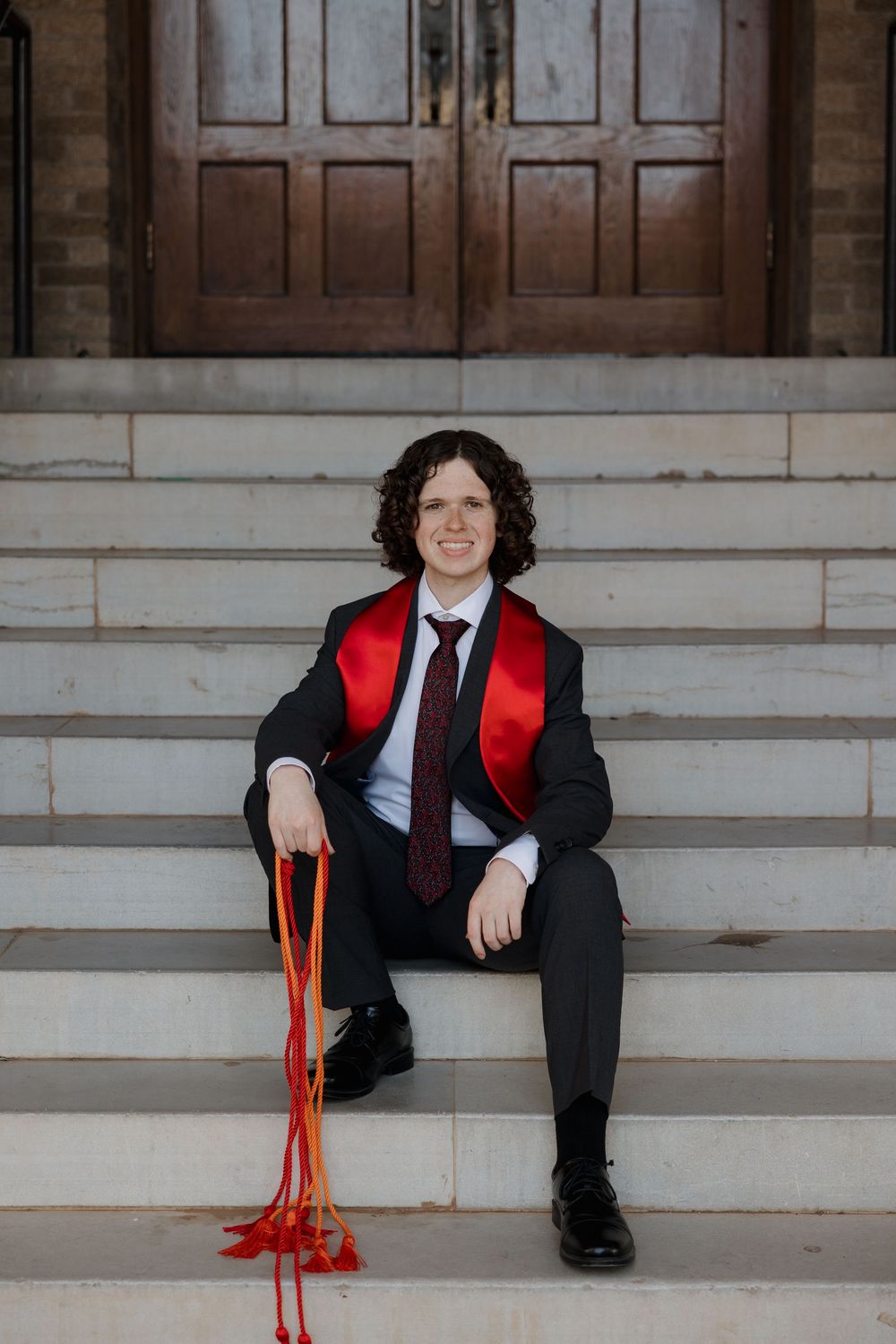 Young man in black suit with red graduation sash sits smiling on stone steps holding orange tassels.