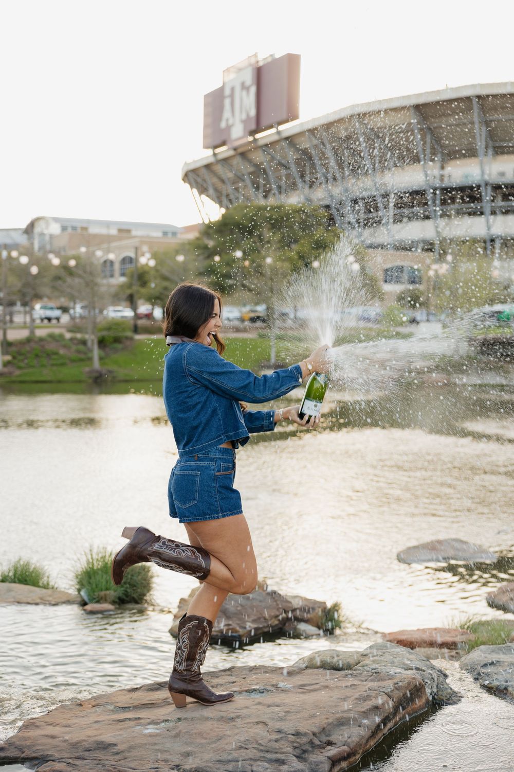 Woman in denim outfit and cowboy boots popping champagne bottle by a pond near a stadium.