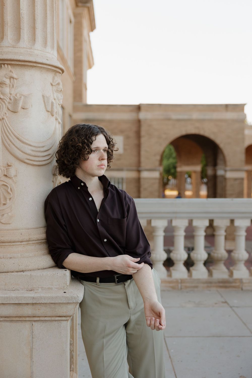 Young man with curly hair in black shirt and khaki pants leaning against ornate stone column at historic building.