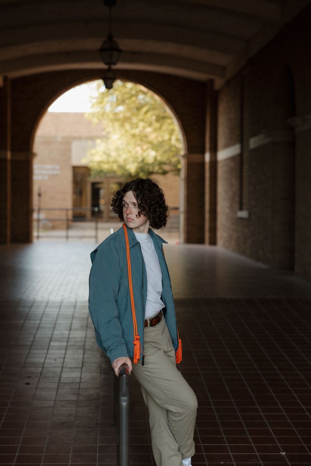 Young person with curly hair in teal jacket and khaki pants leaning on railing under brick archway.