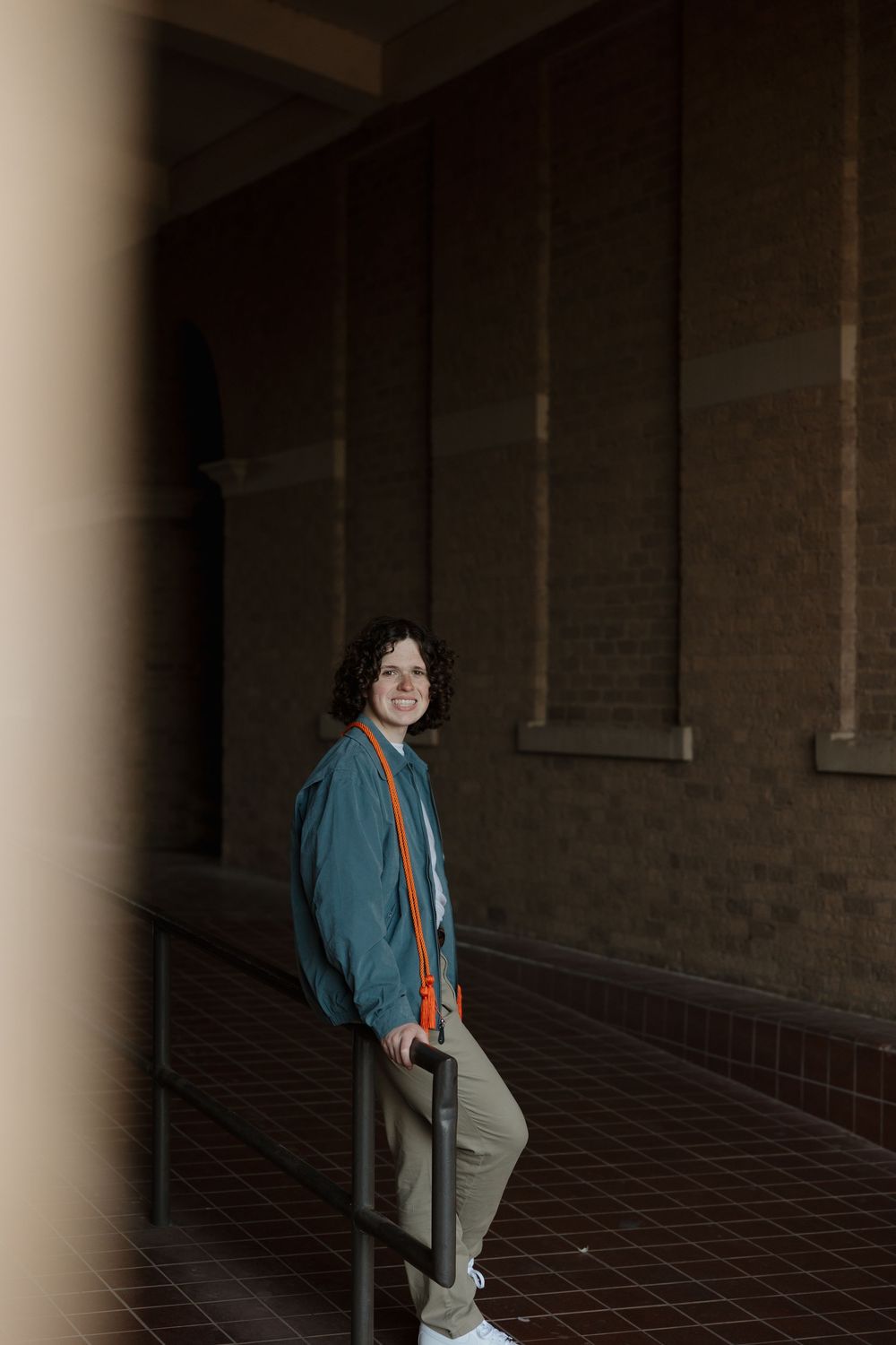 Smiling young person with curly hair in teal jacket and khaki pants leaning on railing in corridor.