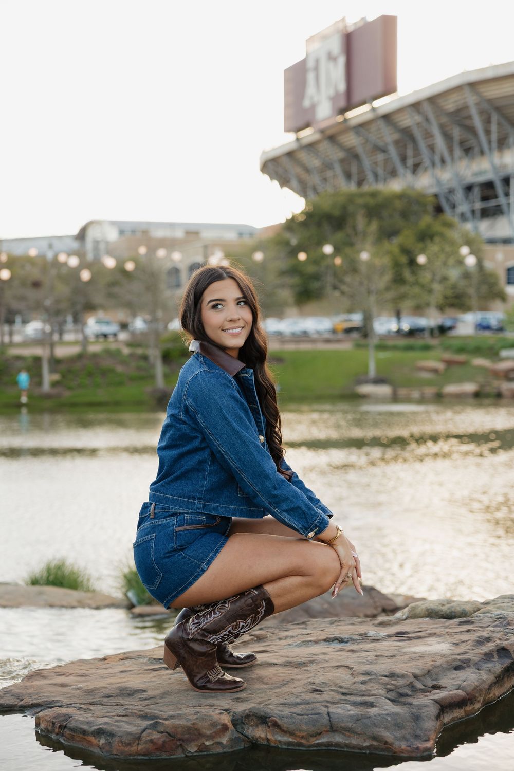 Young woman in denim outfit and cowboy boots poses on rocks near a pond with a stadium in the background.
