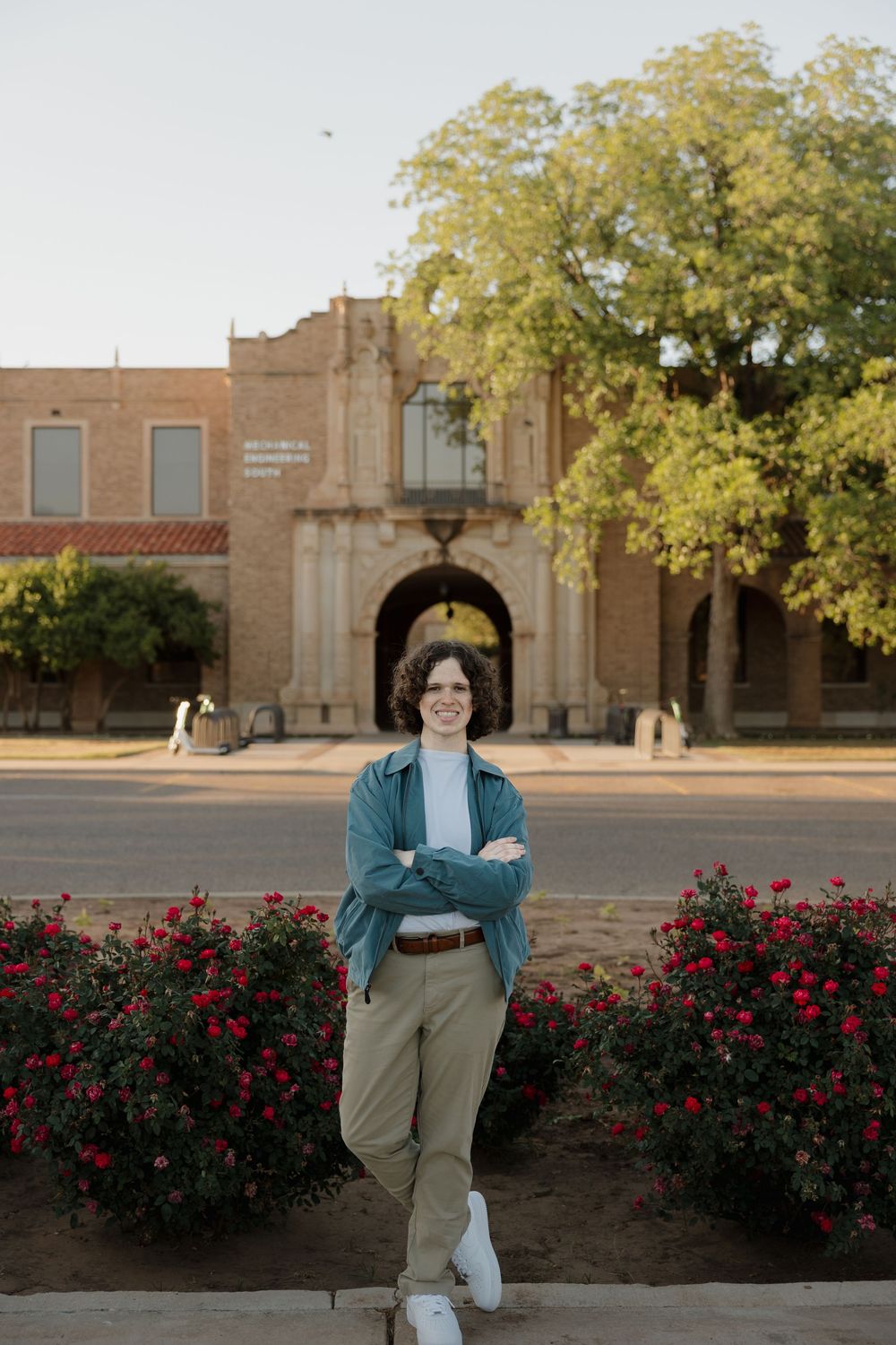 Young person standing arms crossed before a historic building with red roses and green trees in warm sunlight.