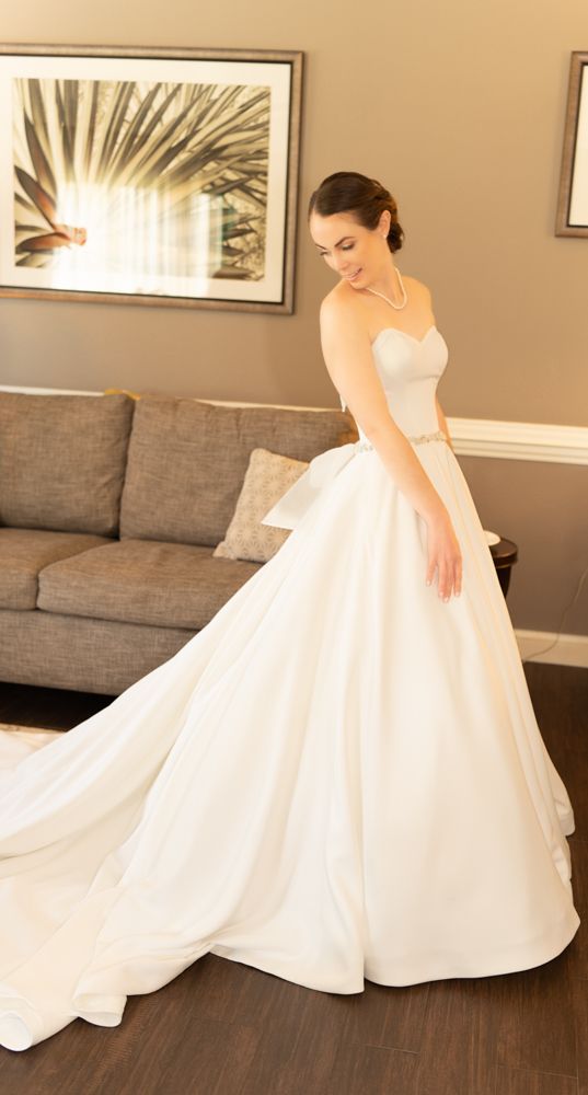 Beautiful strapless white wedding gown with full skirt and train shown in an indoor living room setting with elegant decor.