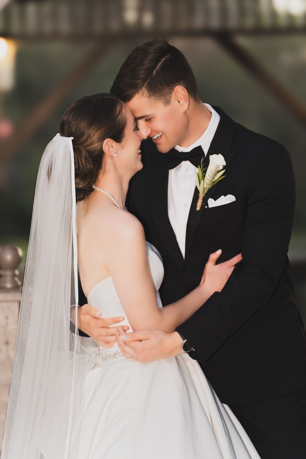 A romantic wedding moment with the bride in white gown and veil embracing her groom in classic black tuxedo.