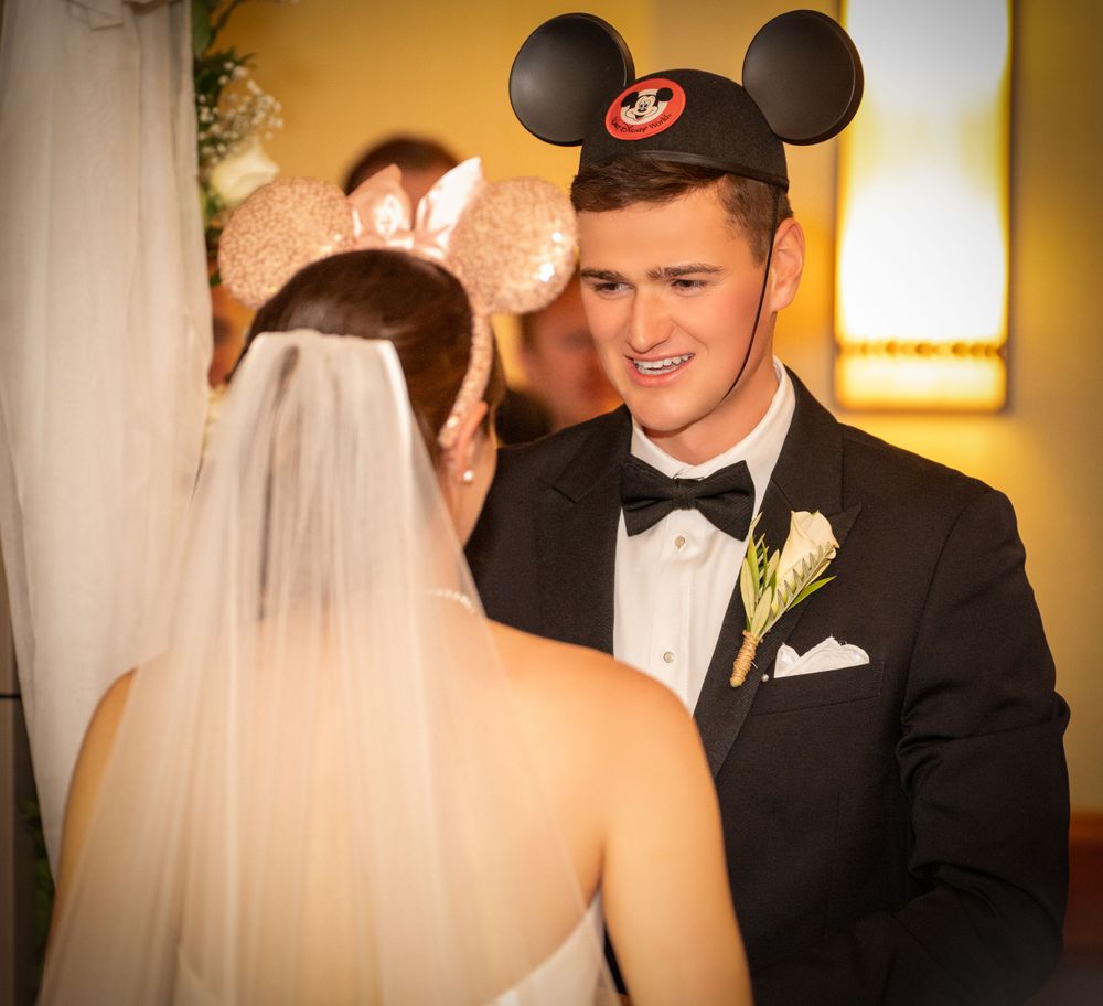 A groom wearing a Mickey Mouse ear hat and tuxedo smiles at his bride during a Disney-themed wedding ceremony.