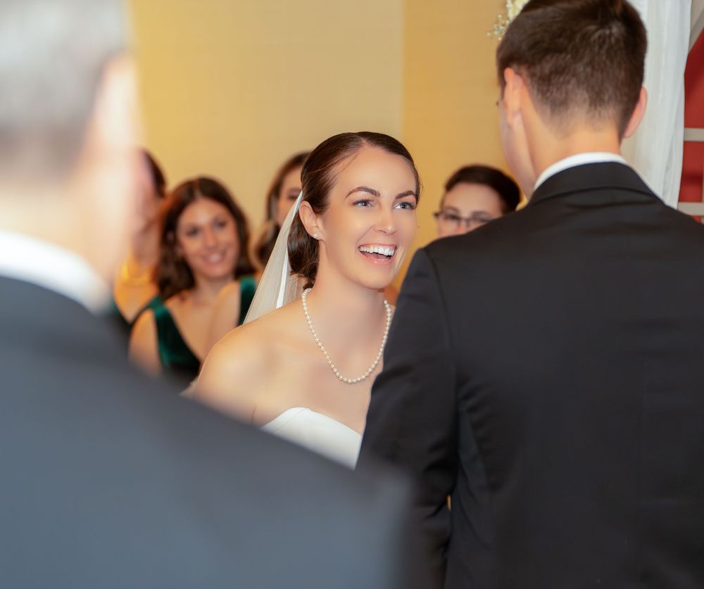A bride in a white wedding dress smiles during a ceremony while exchanging vows in a warmly lit room.