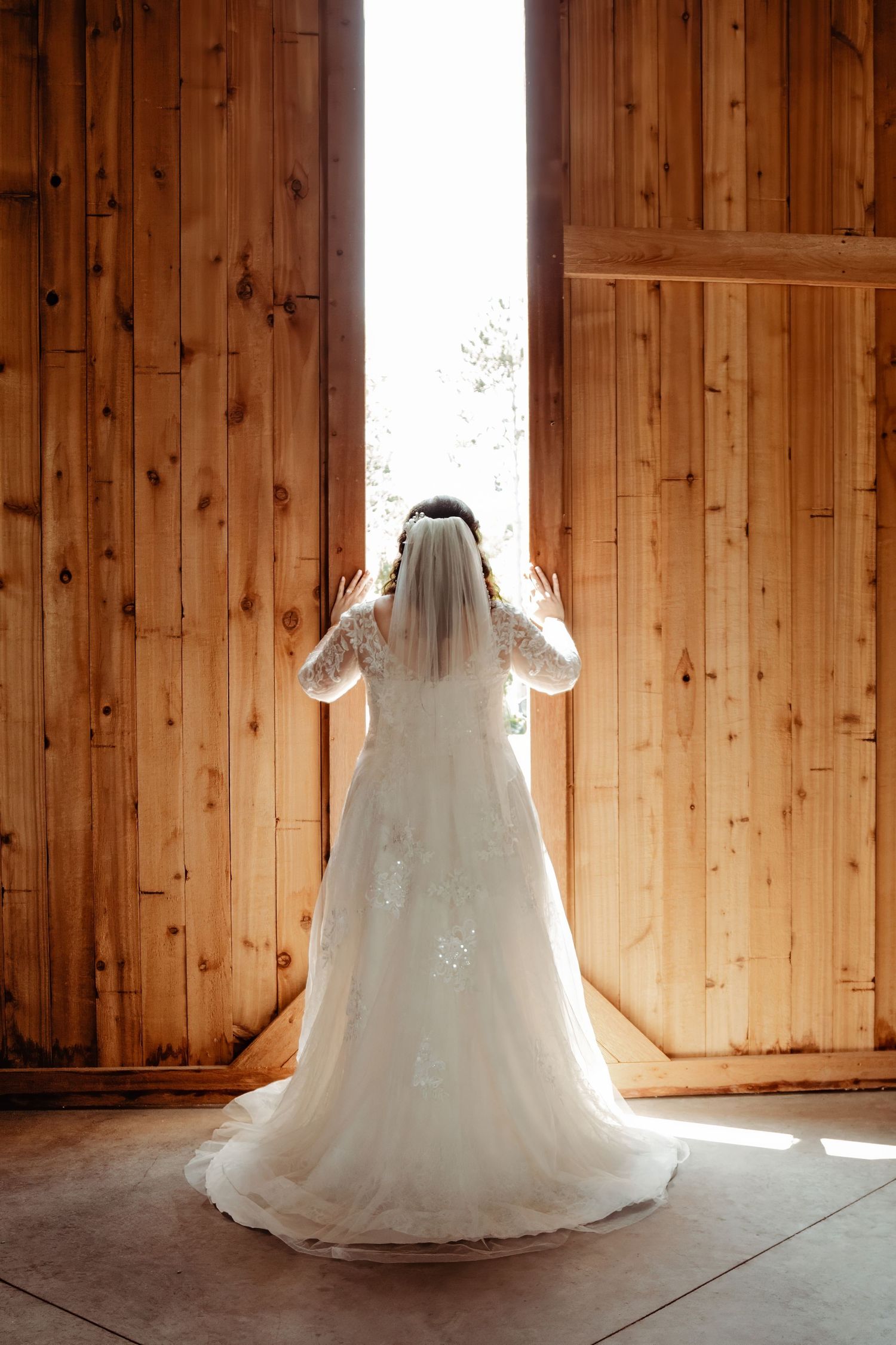 A backlit silhouette of a bride in white lace gown standing between wooden barn doors.