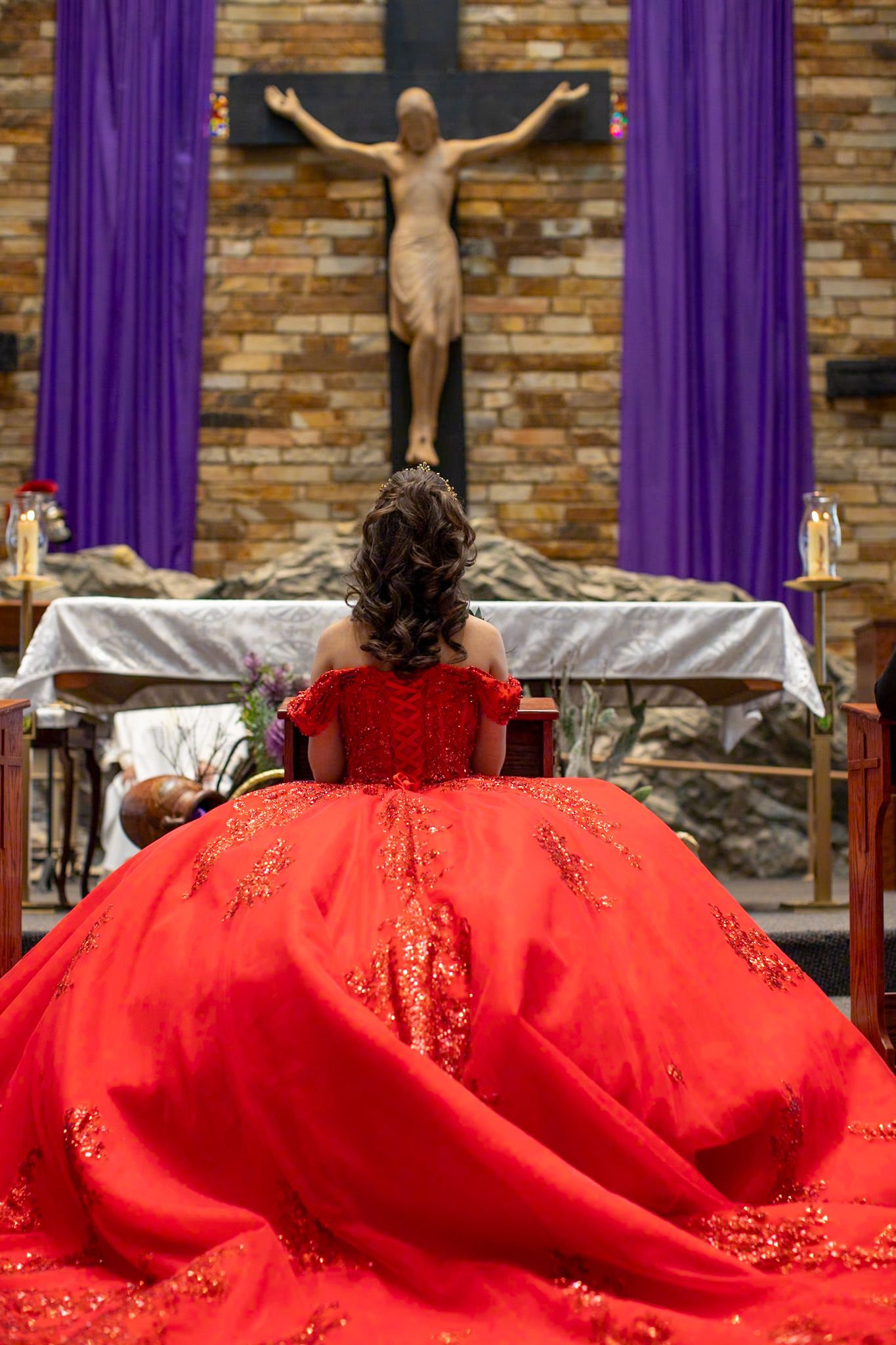 A person in a red embellished gown kneels before a crucifix in a church setting with purple drapes.
