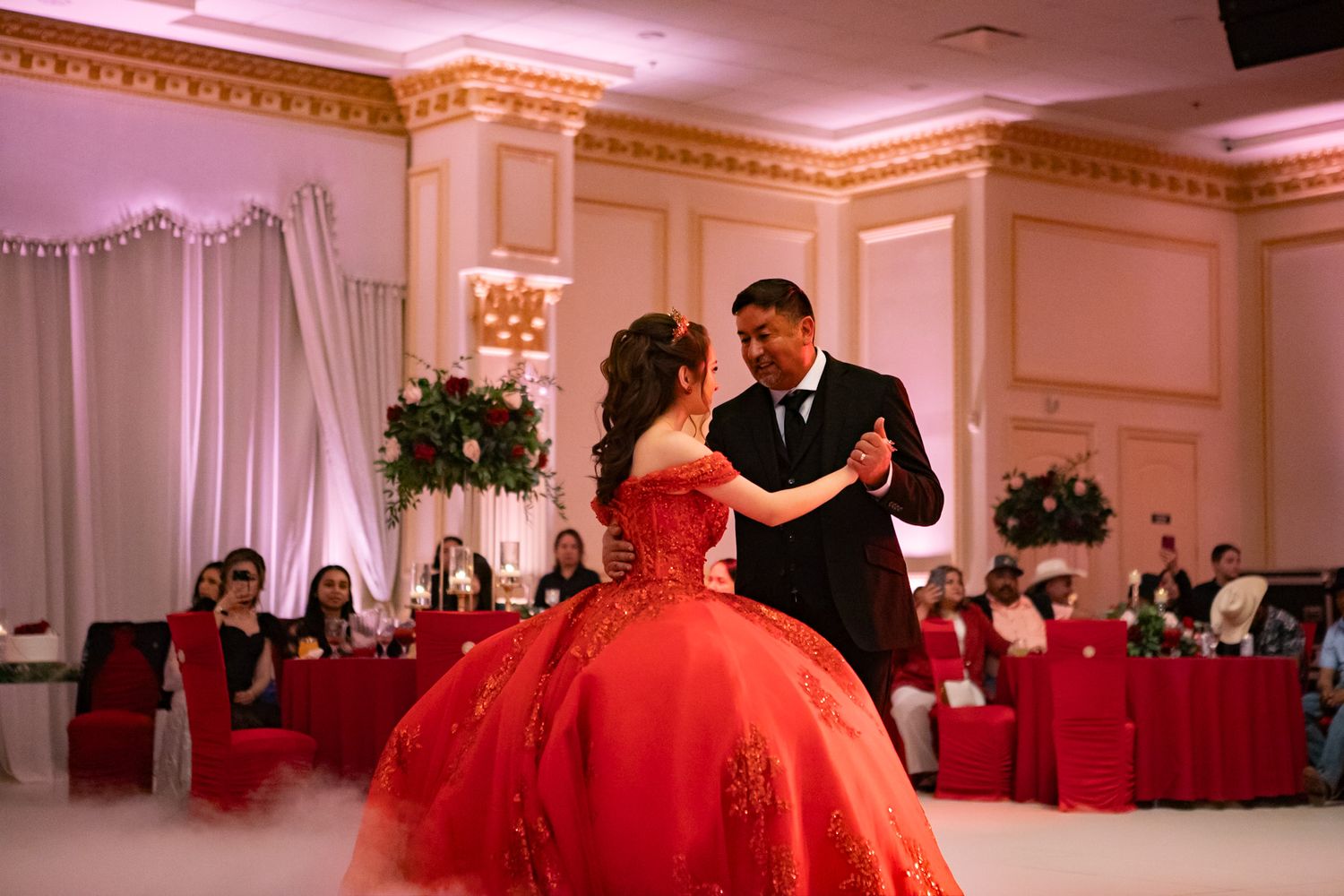 A couple dances in an elegant ballroom with red decor and lighting.