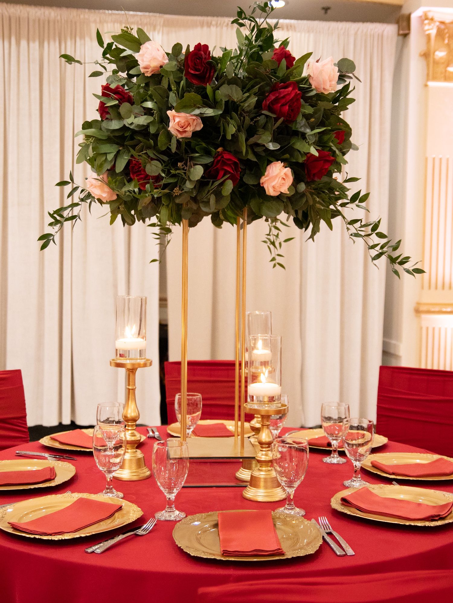 Formal table setting with red tablecloth and tall floral centerpiece featuring red and pink roses.