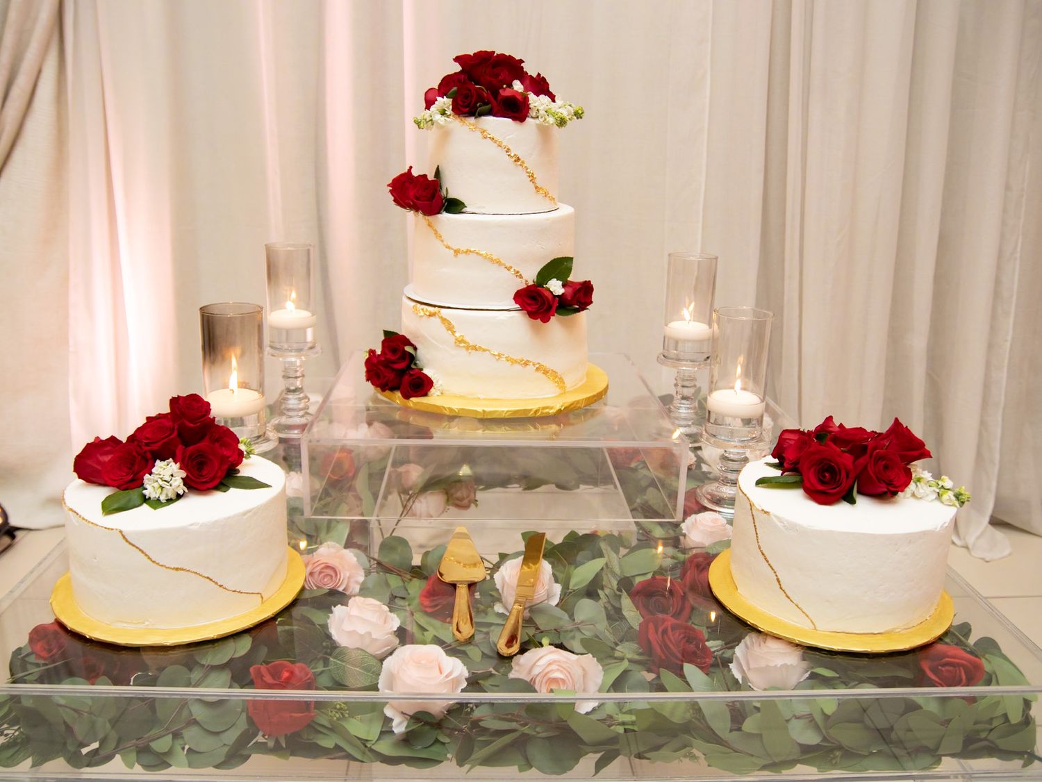 Elegant wedding cake display featuring white tiers with red roses and gold accents on a crystal stand.