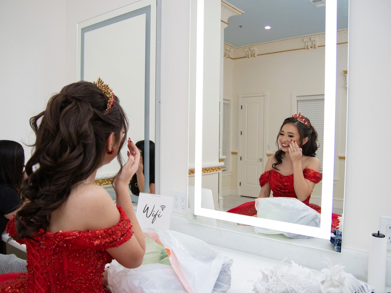 A woman in a red dress gazes at her reflection while getting ready in a brightly lit dressing area.