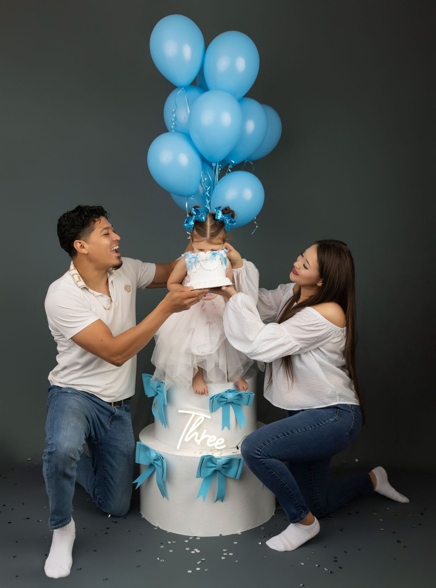 Gender reveal photoshoot with blue balloons and white cake against dark background.
