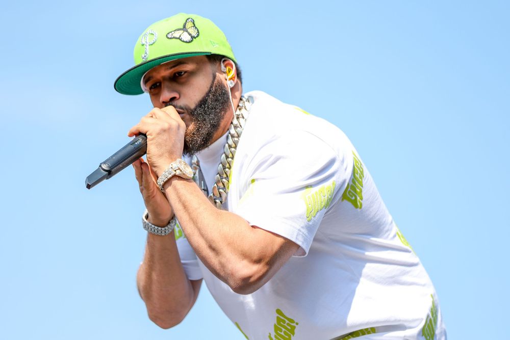 Artist wearing green cap and white shirt performing against blue sky backdrop.