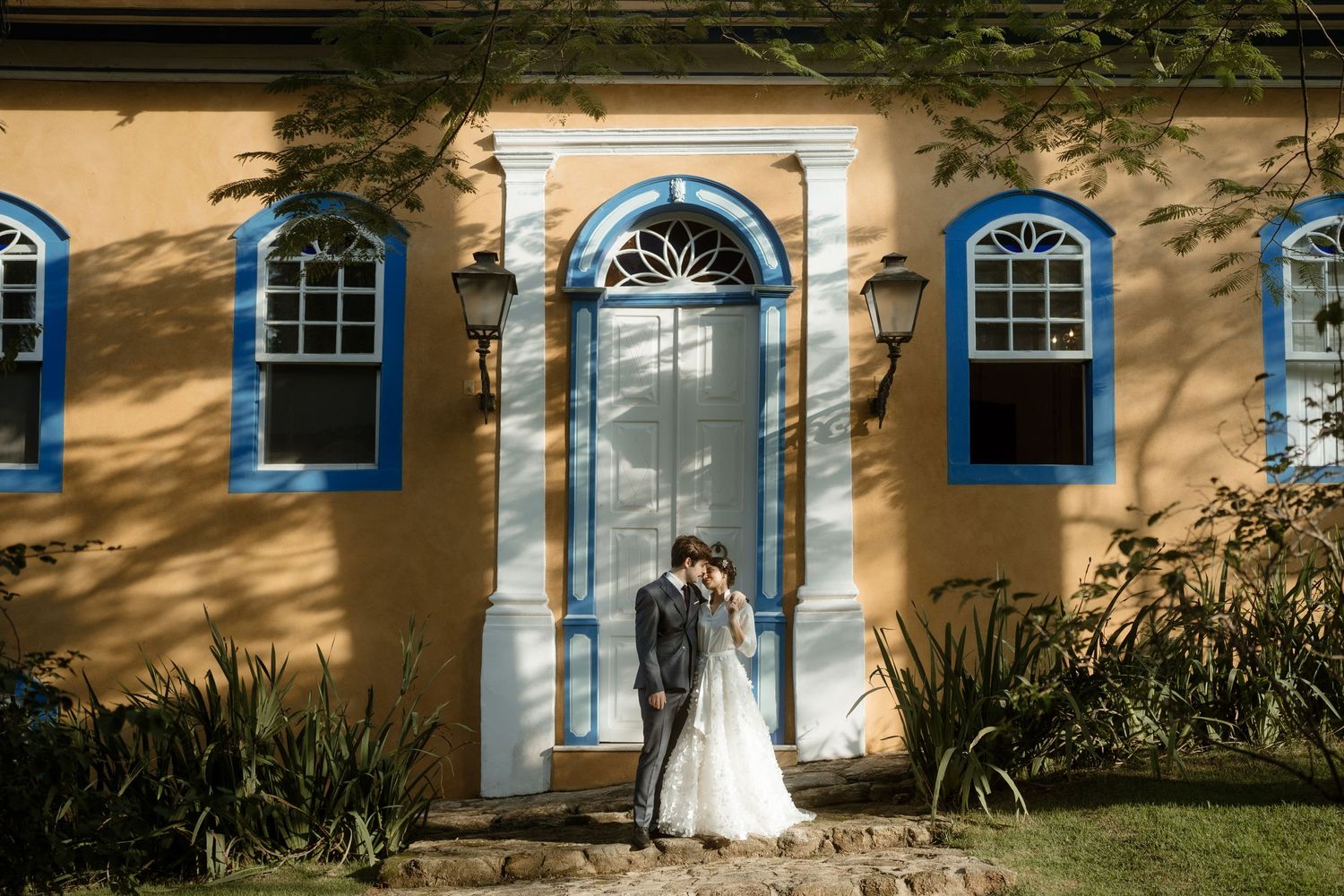 A wedding couple poses in front of a charming yellow building with blue-trimmed windows and door.