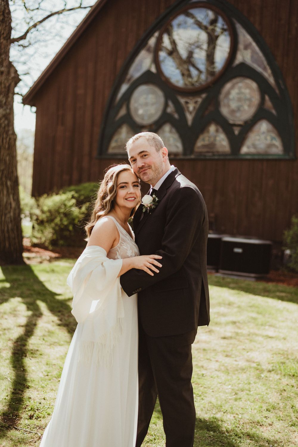Joyful wedding couple embracing in front of rustic chapel on sunny day.