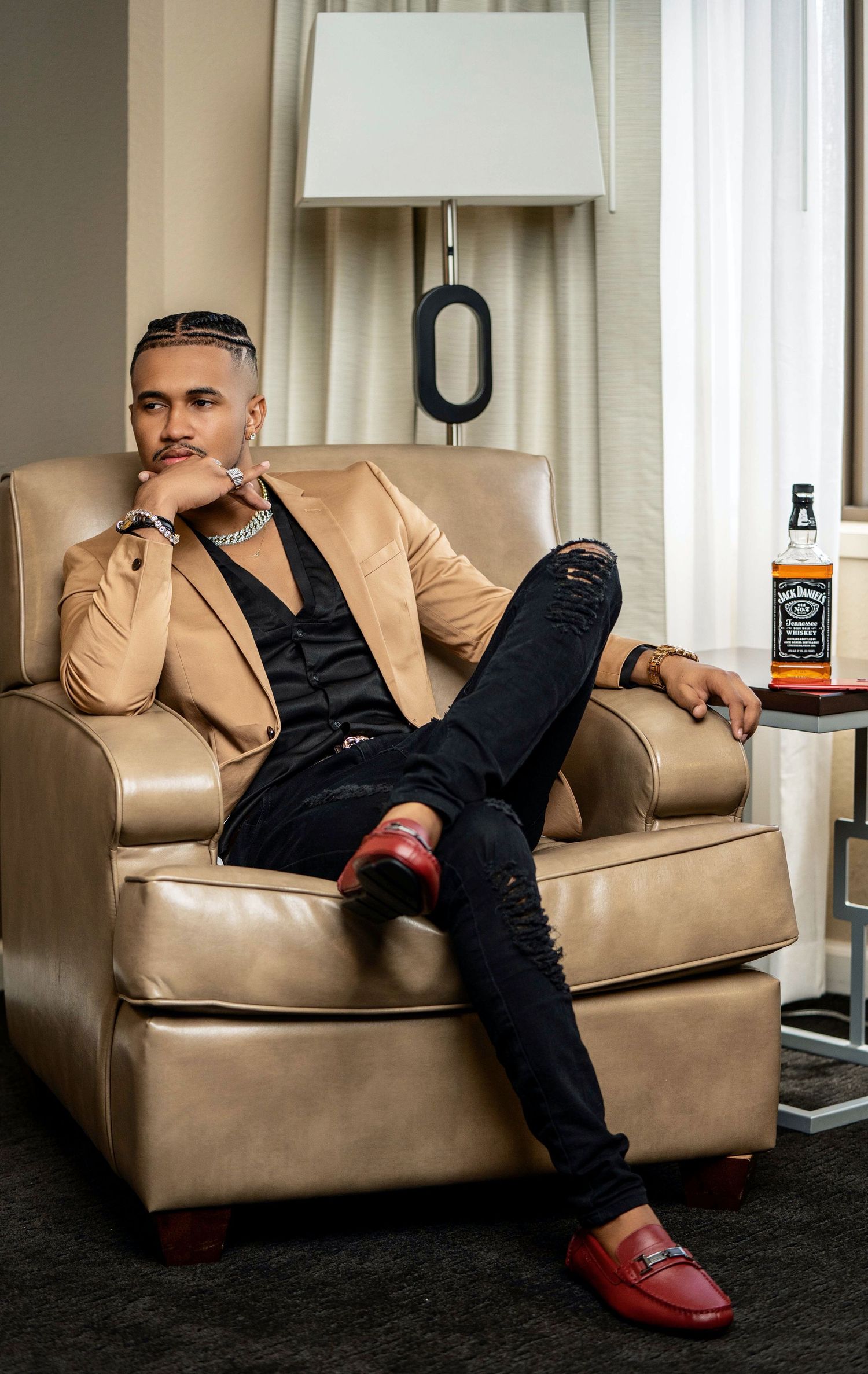 Stylish man in beige blazer and red loafers relaxing in leather chair beside Jack Daniels bottle.