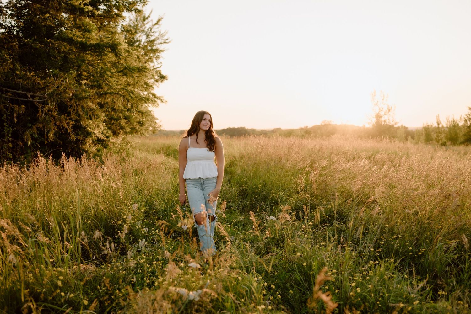 Person walking through tall grass field at sunset wearing casual clothing.