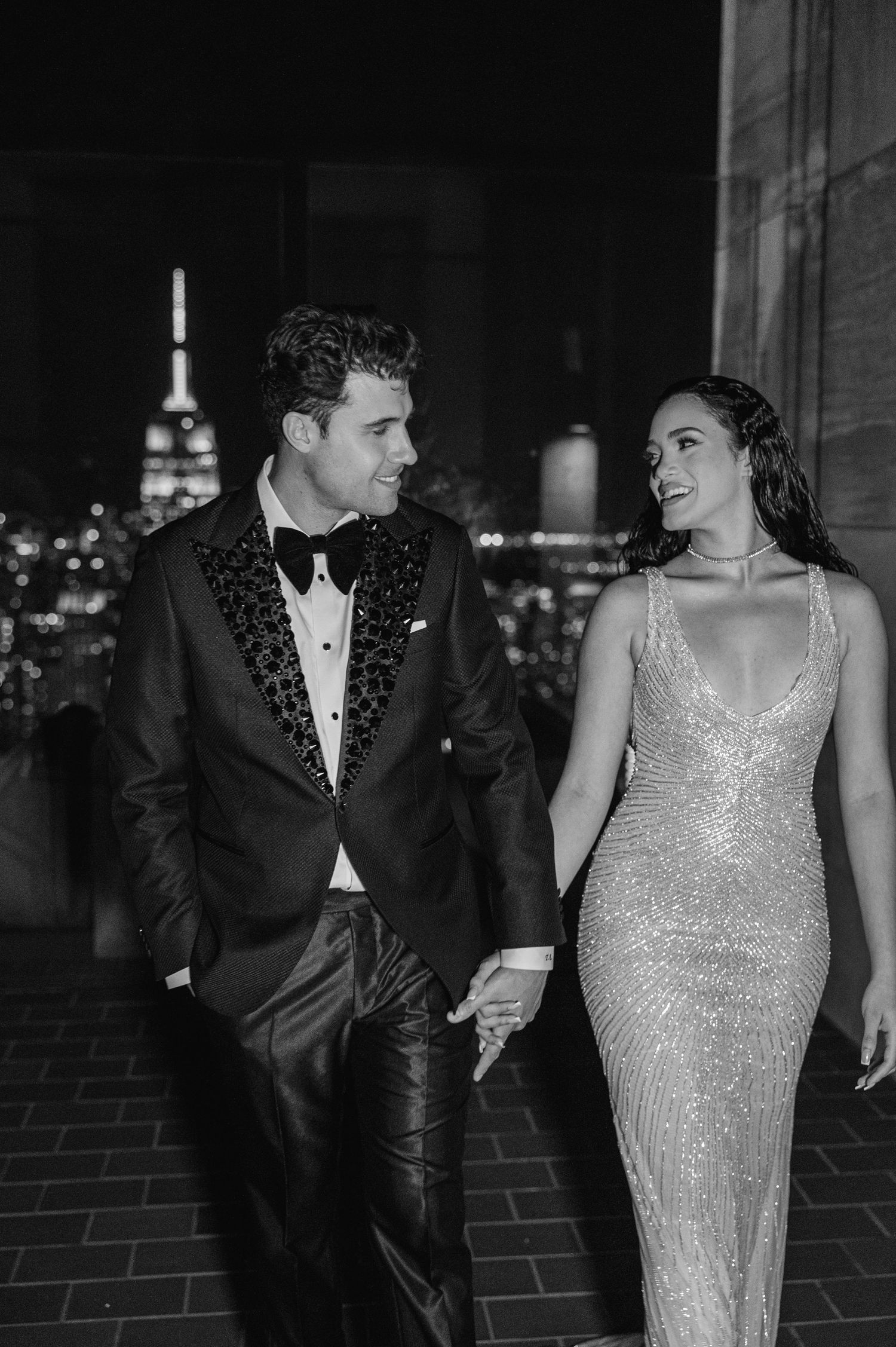 Glamorous couple in formal wear share intimate moment with NYC skyline at night.