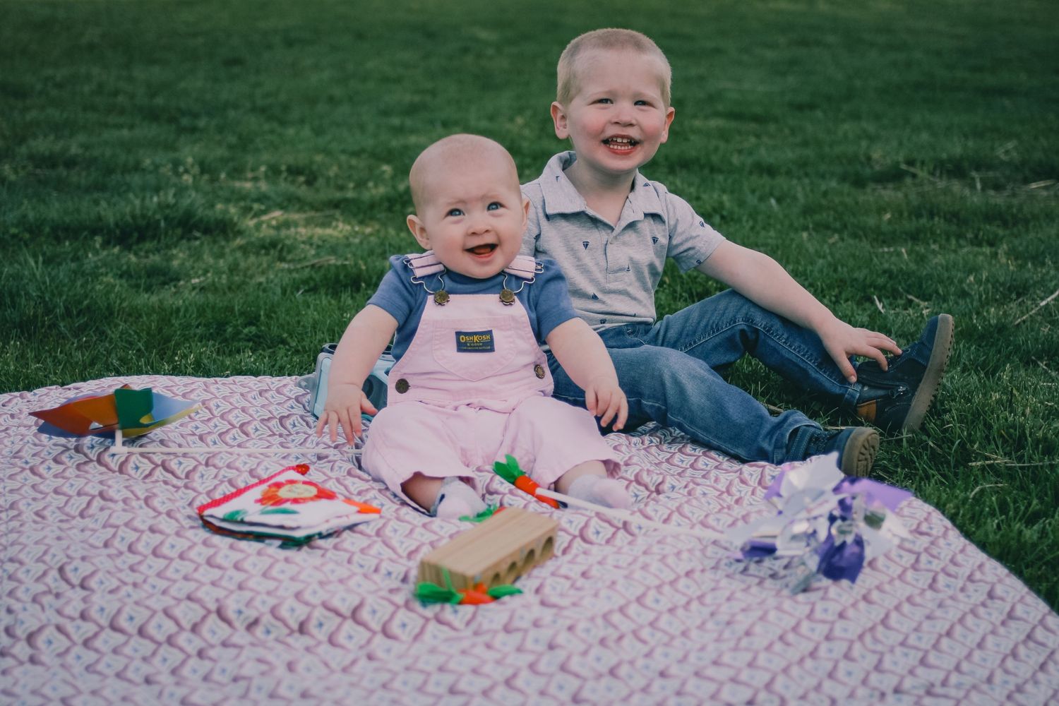 a baby and a toddler sitting on a picnic blanket laughing