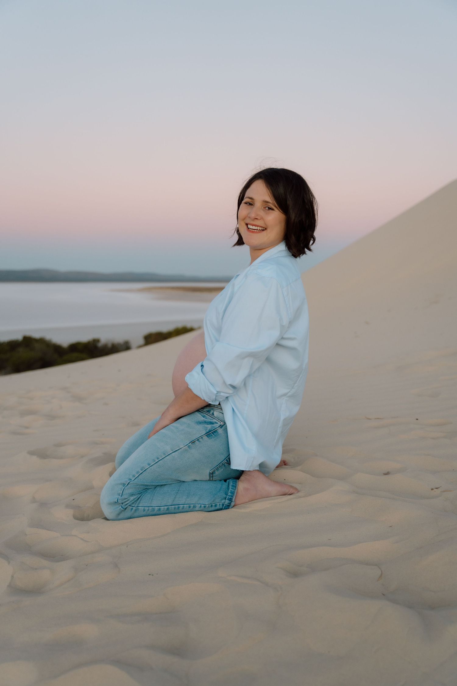 Expectant parent relaxing on sand dunes during golden hour wearing casual denim outfit.