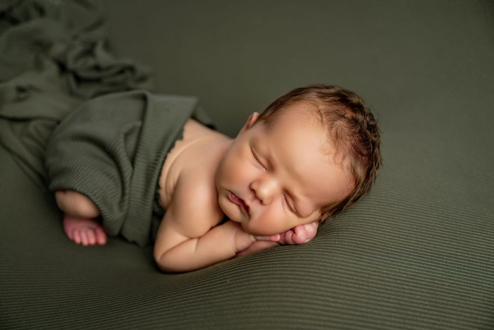 Peaceful newborn baby sleeping wrapped in olive green cloth on matching green backdrop.