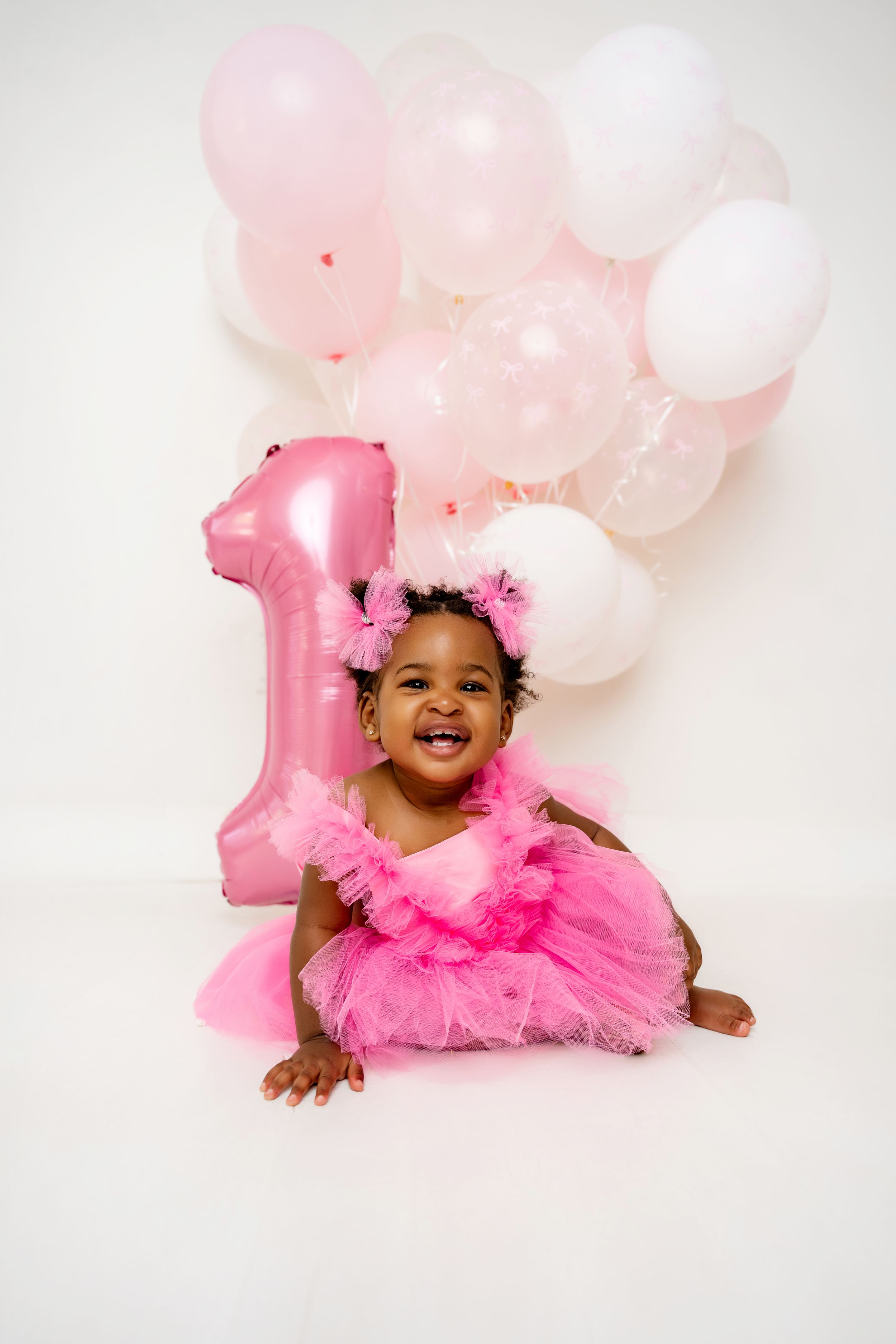 Baby wearing pink tutu dress poses with pink and white balloons against white backdrop for first birthday photoshoot.