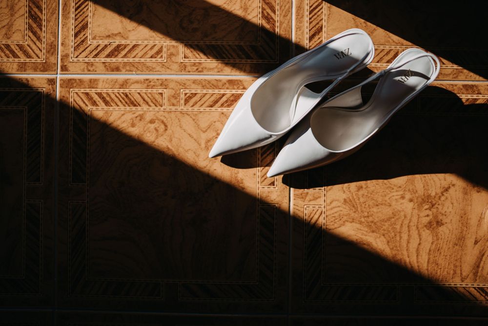 Elegant white bridal shoes on a wooden surface with dramatic diagonal shadows.