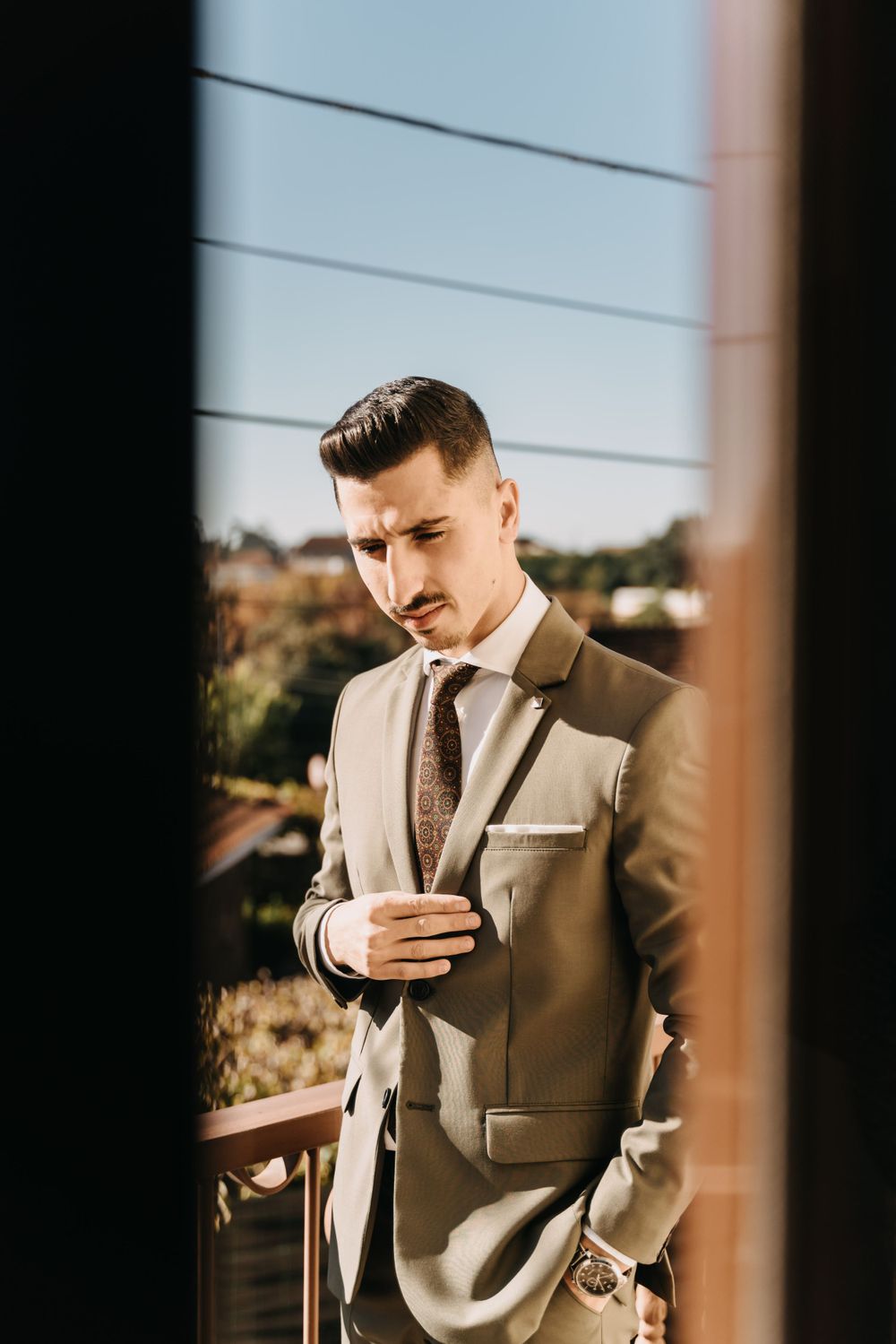 Groom in tan suit adjusts his tie while looking down through window frame.