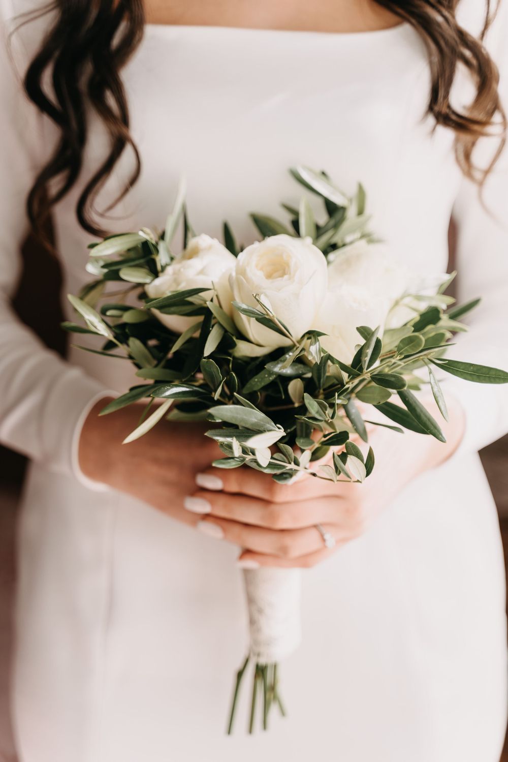 Elegant white rose and olive branch bouquet held against a white dress.