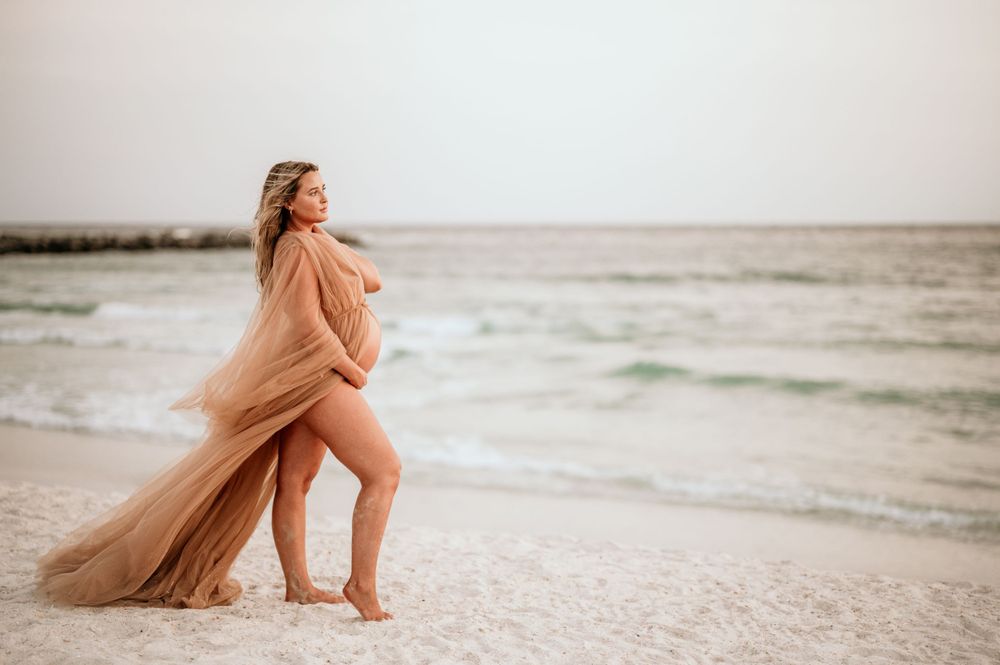 Expectant mother in flowing peach gown poses gracefully on sandy beach at dusk.