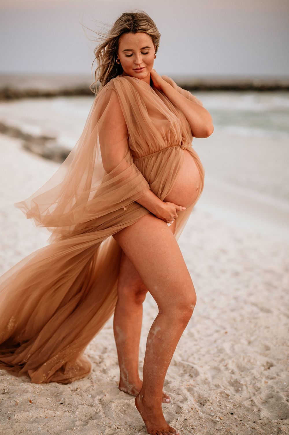 Artistic maternity beach photo with flowing peach dress catching the coastal breeze.