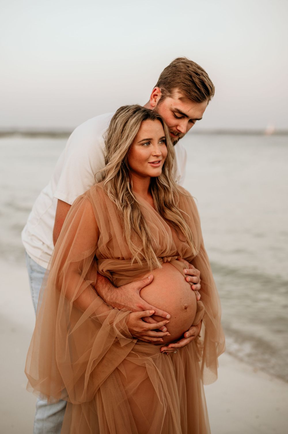 A maternity photo session at the beach featuring a flowing peach-colored dress and intimate couple pose at sunset.