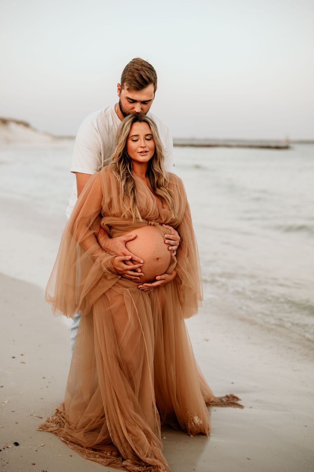Romantic beachside maternity photoshoot with couple embracing in a flowy tan dress.