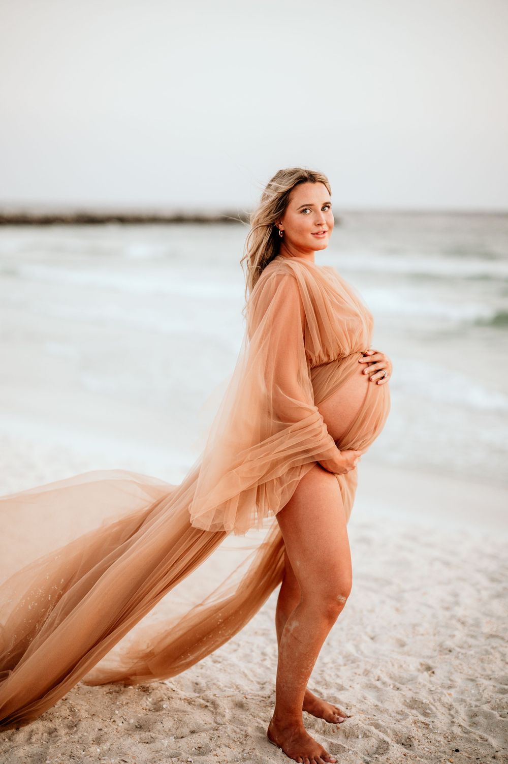 Expectant mother in flowing peach dress poses gracefully on sandy beach during photo session.