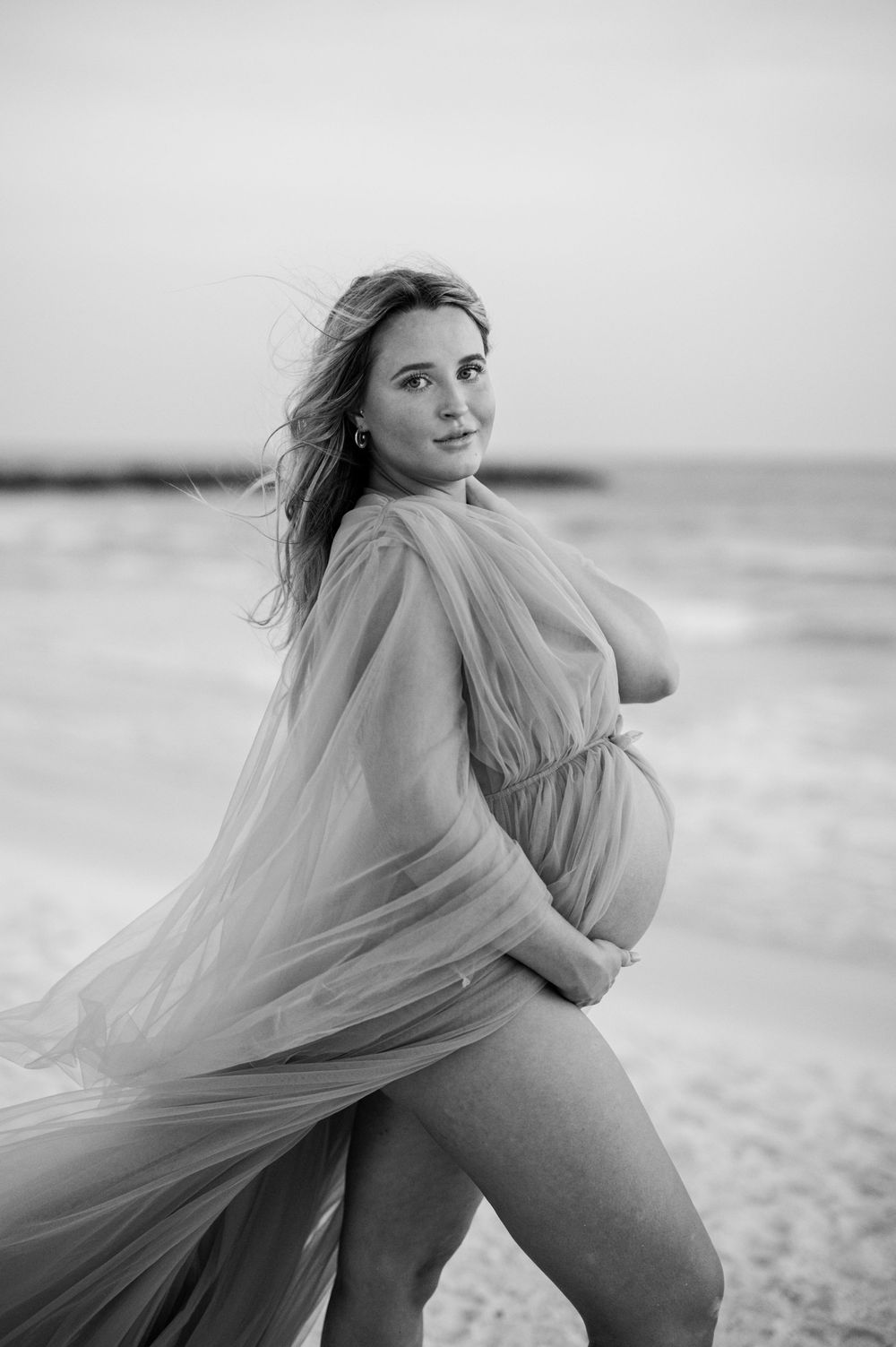 Black and white maternity portrait on beach with flowing dress and natural lighting.