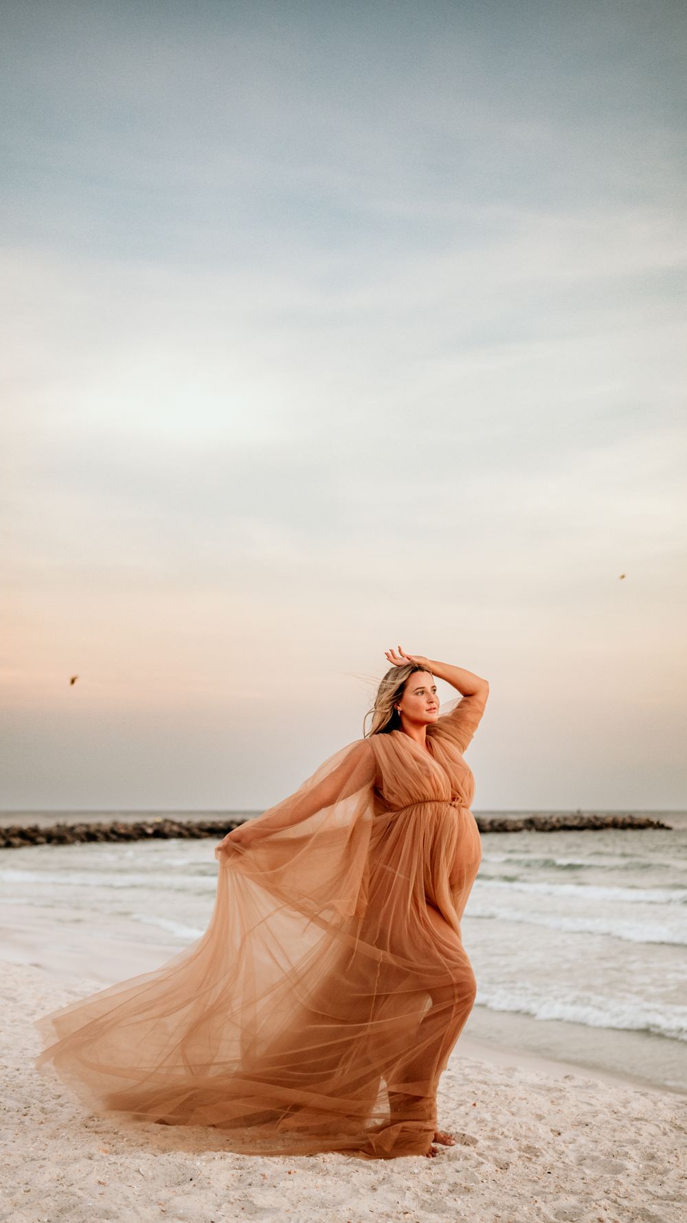 Dramatic beachside maternity portrait with billowing peach dress at dusk.